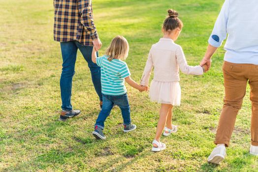 Rear side view of two children walking hand in hand with their dads in the park in a sunny day.