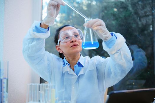 African American woman, scientist chemist dripping few reagent into a flat-bottomed flask with a blue solution and analyzing the going on chemical reaction while conducting experiment in a biolab