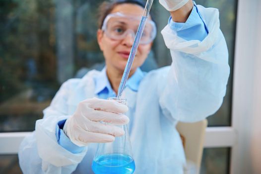 Focus on laboratory glass pipette in the hands of blurred woman scientist, medical biologist, researcher in a white lab coat, safety goggles and gloves pipetting blue liquid into a flat-bottomed flask