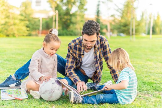Front view of a young father with his children sitting on the grass in a park reading a children's book. Concept of single parent family.