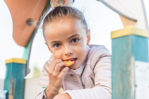 Portrait of a little girl looking at camera eating a cookie in a playground.