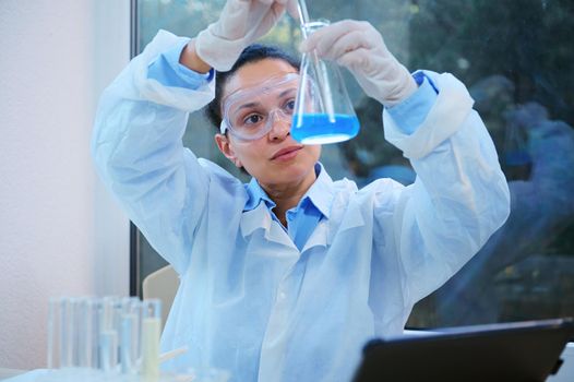 Multi-ethnic woman scientist, in lab coat and safety eyeglasses using a glass stick mixing the blue solution in a flat bottomed flask, while conducting a chemical experiment in a science laboratory