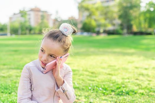 Front view of a little girl looking away talking on phone in the park with copy space.