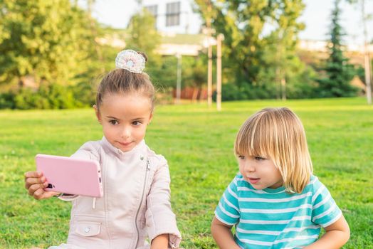 Front view of a a little girl taking a selfie with a phone with her little brother in the park