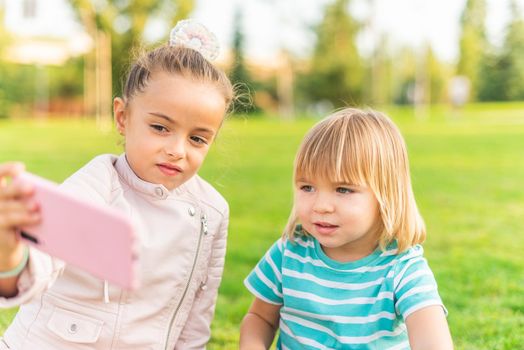 Portrait of sister and brother sitting on grass taking a selfie in the park in a sunny day.