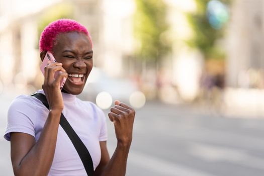 African young woman with pink short hair laughing while talking to the mobile in the streeet