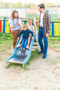Front view of a gay male couple with their adopted son teaching him how to play on the slide at a playground.