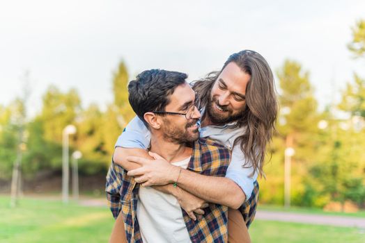 Front view of a young gay male couple having a good time in the park while one man carries the other on his back.