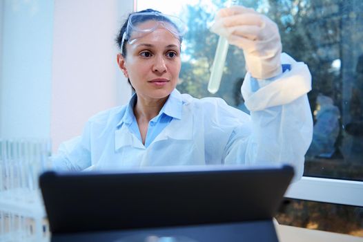 Confident experienced multi-ethnic woman in lab wear, scientist holding a test tube and analyzing the chemical reaction going on, sitting in front of a digital tablet in a chemistry science laboratory
