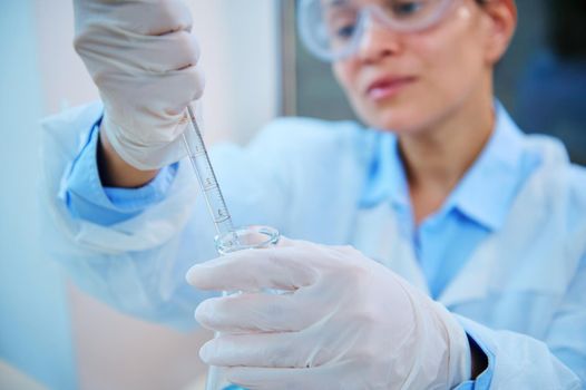 Focus on glass laboratory pipette in gloved hands of blurred female pharmacologist, medical biologist, scientist chemist dripping few drops of chemical reagent into flask while conducting experiment