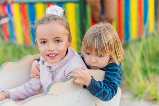 Portrait of a happy little sister and brother sitting in a game of a playground looking at camera.