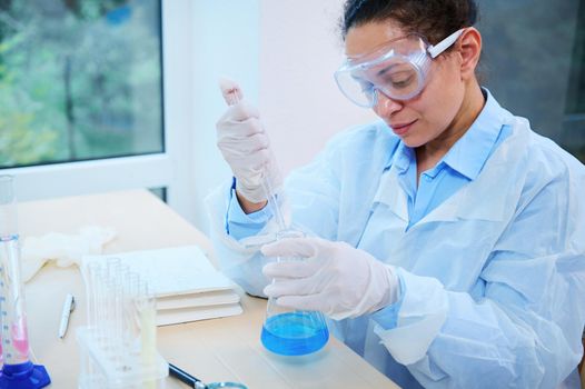 African American woman, scientist pharmacologist pipetting the blue chemical liquid in a flat-bottomed flask, sitting at a table in a light laboratory interior, conducting a science experiment