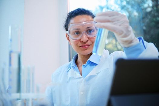 Close-up of beautiful African American woman, scientist, pharmacologist, clinical researcher wearing safety goggles, examining test tubes with chemical substances, in a science biological laboratory