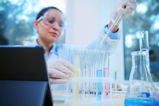 Selective focus on a glass pipette with dripping chemical reagent into test tubes over blurred female scientist pharmacologist, medical biologist working on vaccine in scientific research laboratory