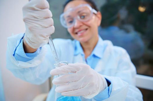 Selective focus on glass dropper in gloved hands of blurred woman scientist chemist wearing safety goggles and lab coat, doing science experiments, pipetting chemical solution in biological laboratory