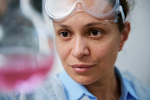 Headshot of confident experienced multi-ethnic woman, scientist chemist, pharmacologist inspecting the chemical reaction taking place in a glass flask with pink chemical fluid substance in laboratory