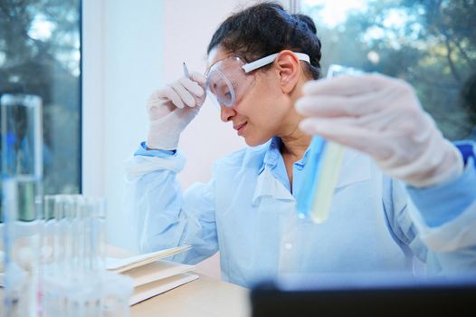 Experienced middle-aged multi-ethnic woman scientist chemist, pharmacologist holds a test tube with blue liquid substance and using tablet for medical research, works in chemical science laboratory.