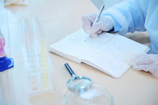 Cropped view of a scientist with gloved hands, sitting at a table with a magnifying glass and lab glassware, holding a pen and writing on a notepad the results of conduct experiments in laboratory