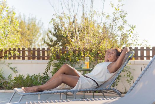 Full length view of an adult woman relaxing on a lounge chair in a country hotel on vacation with an orange juice next to her.