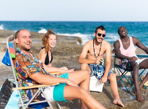 four young people sunbathing on a rocky beach seated on chairs, horizontal close up