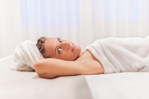 An adult woman wrapped in towels after showering lying in bed and looking at camera with her arms under her head and her eyes closed. Concept of hotel.