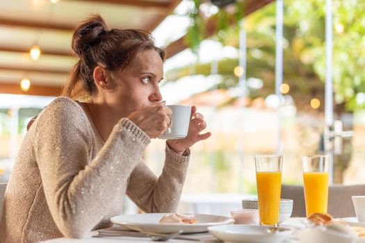 Side view of an adult woman having breakfast in a hotel dinning room while drinking a coffee and looking away. Concept of breakfast in hotel.