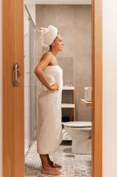 Full side view of an adult woman wrapped with towels looking at mirror with her hands on her waist in a bathroom of a hotel. Concept of hotel.