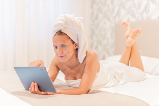 An adult woman wrapped in towels using her digital tablet while lying down on the bed of a hotel bedroom. Concept of vacation, hotel.