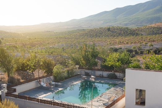 View of a rural house with a swimming pool and a woman relaxing on a deck chair with a landscape of hills and mountains in Almeria, Spain.