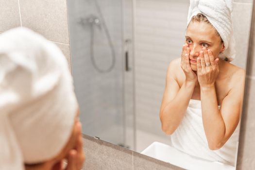 An adult woman looking at herself in the mirror after taking a bath, holding her face with her hands and worrying about the passage of time and wrinkles. Concept of aging.