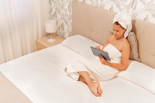 High angle view of a woman wrapped in towels using her digital tablet while lying on the bed of the bedroom of a hotel . Concept of vacation, hotel.