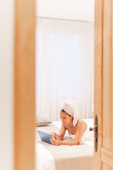 View through the door of a woman wrapped in towels using her digital tablet while lying down on the bed of the bedroom of a hotel. Concept of vacation, hotel.