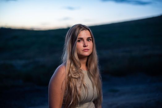 Portrait of a blonde woman looking at the camera with serious expression outdoors as it gets dark.