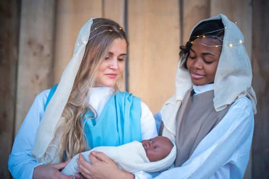 Lesbian caucasian and afro woman representing virgin mary while holding a black baby Jesus in a crib