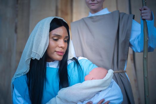 Transgender woman representing Virgin Mary embracing Jesus baby in a christian crib next to Joseph