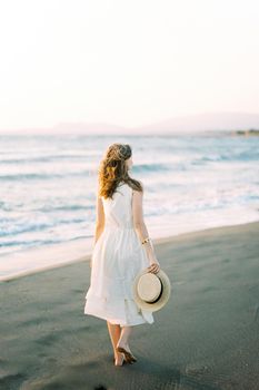 Young woman in a dress barefoot with a hat in her hands walks along the beach. High quality photo