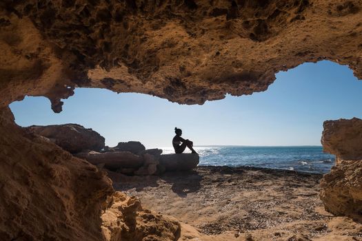 Young woman sitting next to a cave of a rock formation in front of the sea in Formentera in Spain.