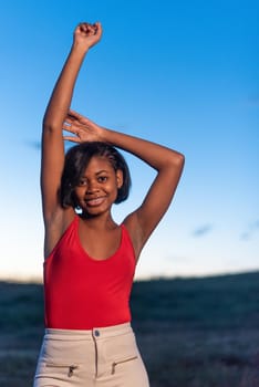 Vertical portrait of a young black woman raising her arms while posing during dusk outdoors