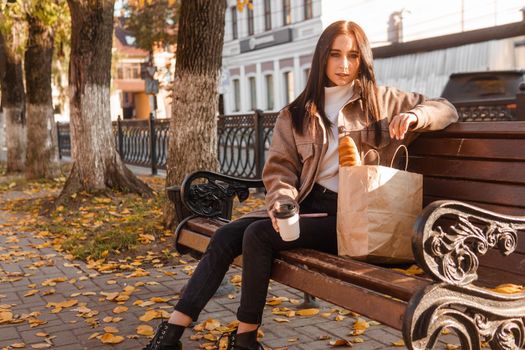A stylish brunette woman walks around the autumn city. The brunette is sitting on a bench outside with a glass of coffee and a phone, holding a craft bag with a baguette.