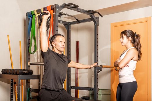 Athlete man doing shoulder press exercises with kettlebell while woman look at him in gym. Concept of exercises with equipment in gym.