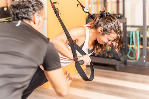Sportswoman doing exercise with trx fitness straps in gym with help of trainer. Concept of exercises with equipment in gym.