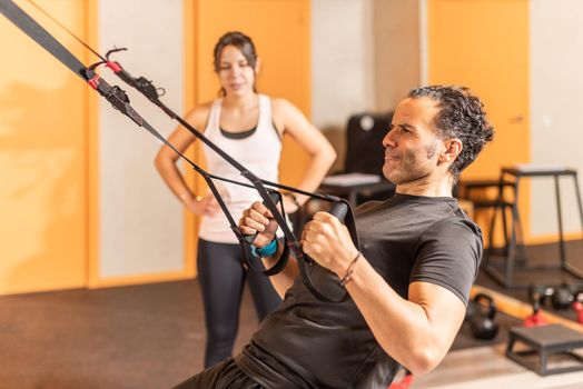 Sportsman doing exercise with trx fitness straps while woman look at him in gym. Concept of trx training.