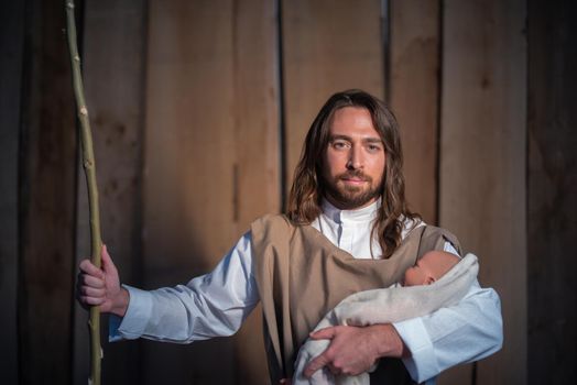 Man representing the biblical character of Joseph facing the camera and holding Jesus baby in a crib