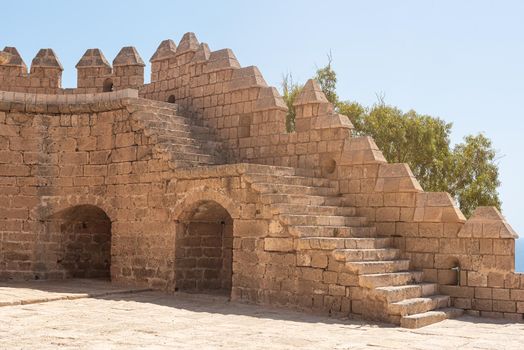 Exterior of ancient stone walls and arched passage under staircase of historic construction of castle, and fortress of Citadel of Almeria city in Spain in summer sunny day