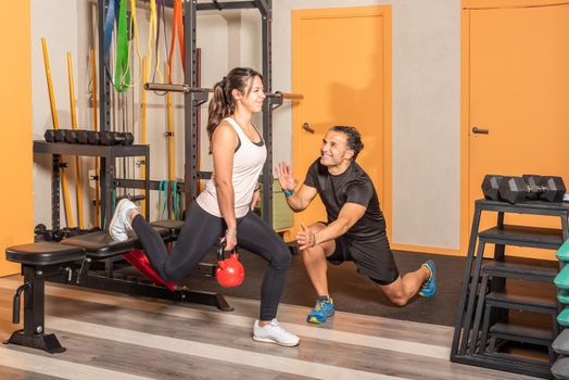 Sportswoman in sportswear doing lunges with a kettlebell in her hand with one leg resting on a bench with help of coach in gym. Concept of exercise with equipment in gym.