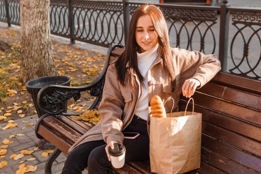 A stylish brunette woman walks around the autumn city. The brunette is sitting on a bench outside with a glass of coffee and a phone, holding a craft bag with a baguette.