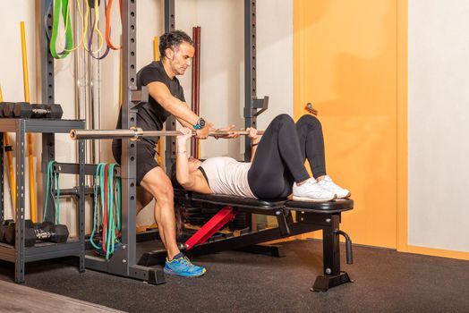 Sportswoman exercising on bar bench press in a gym with help of coach. Concept of exercises with equipment in gym.