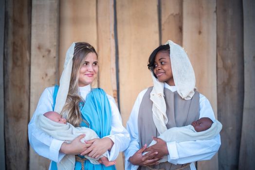 Multiethnic lesbian woman representing Virgin mary looking each other and holding a baby in a crib