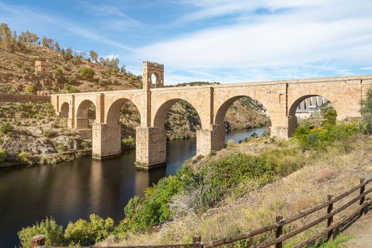 Roman stone bridge over calm river and green trees with bushes on mountainous shores in daylight in Spain