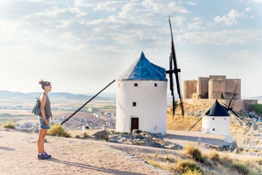 Woman in summer clothes, sunglasses and bag standing standing next to some ancient windmills an a castle in Consuegra, Spain.